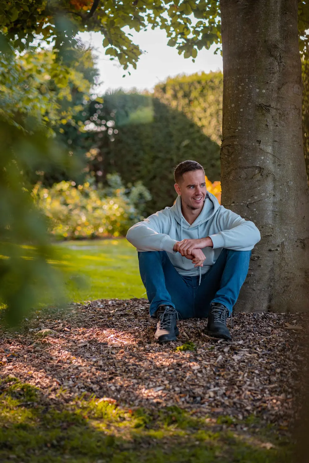 Jochem zit ontspannen tegen een boomstam in een park, armen over zijn knieën