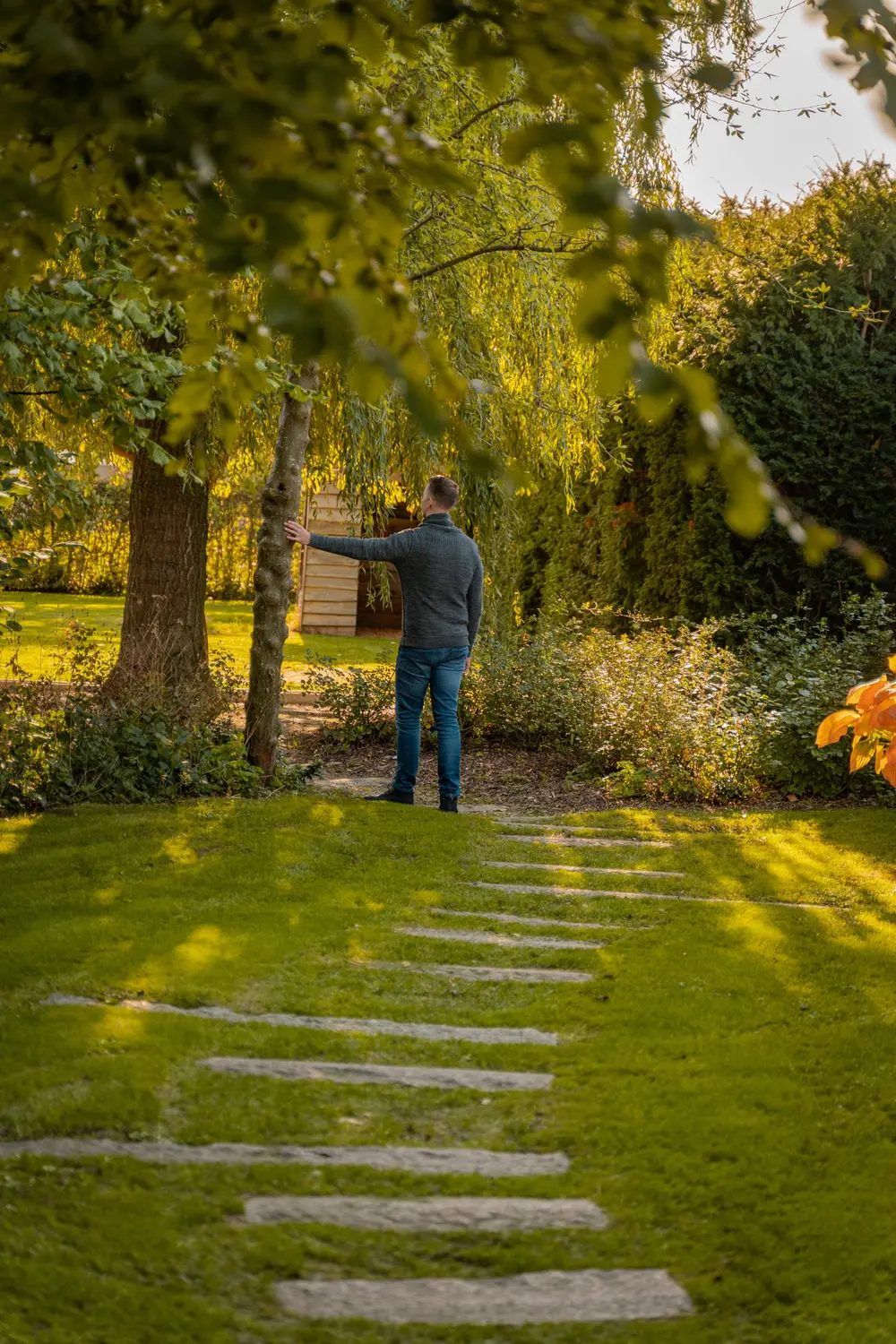 Jochem staat met zijn rug naar de camera in een groene tuin, zijn hand tegen een smalle boom